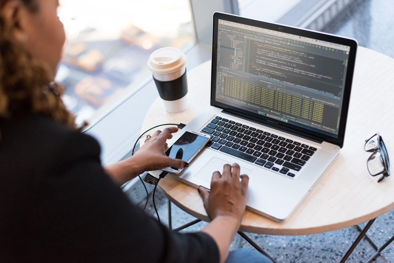 Person sitting at a small table with a laptop, coffee, phone, and glasses in front of them.