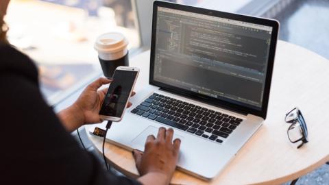 Black woman sitting at a small table writing computer code. She holds a tethered phone in her hands. Also on the table are a cup of coffee and glasses.